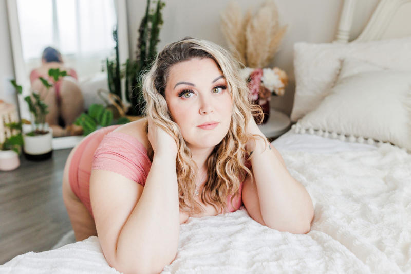 Boudoir Photograph of a beautiful curvy blonde woman kneeling on the floor while she leans onto a bed with flowers and a mirror in the background. Taken at Meaghan Harvey Photography's Nanaimo Boudoir Studio.