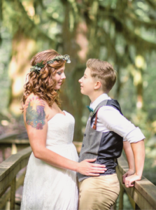 Lesbian couple embrace while standing on a bridge in the middle of a forest after their intimate wedding.