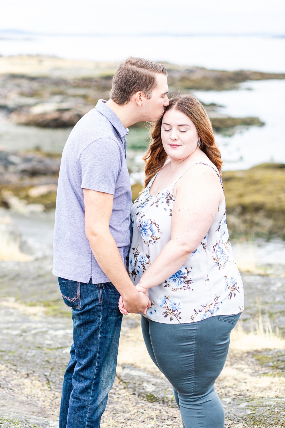 Man kisses his fiancees forehead while they pose for engagement photos at cattle point on Vancouver island